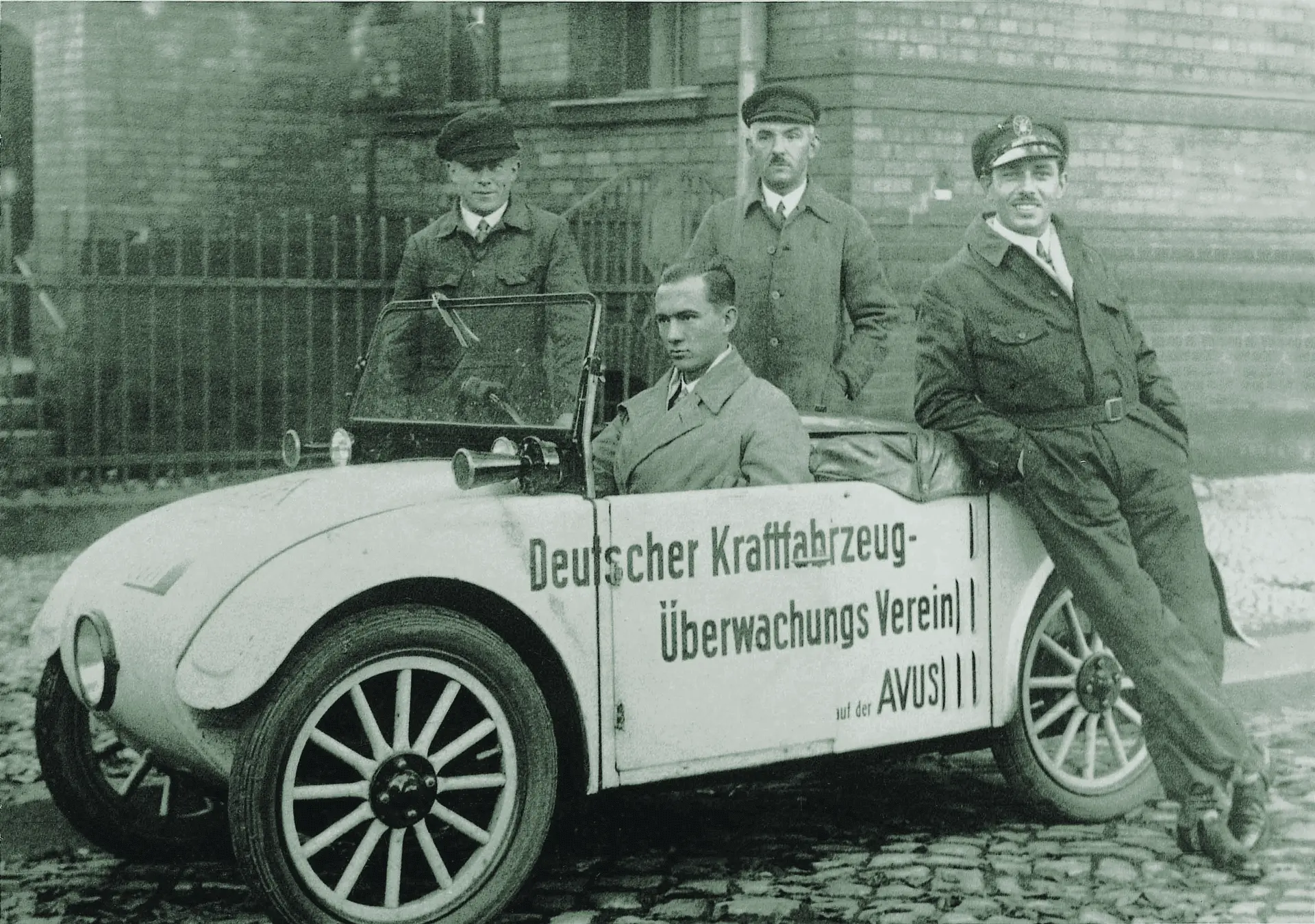 Mustavalkoinen kuva vanhasta autosta, jonka kuskin paikalla on mies ja jonka vieressä seisoo kolme miestä nojaamassa autoon.